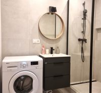 A bathroom with a washing machine, shower cabin, and round mirror in a 2-room apartment.