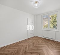 A room in a three-room apartment with a wood-patterned floor and a large window.