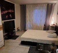 A studio apartment with gray-white walls, a bed, a television, and tiled flooring.