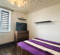Bedroom in a 4-room apartment with a bed, television, wood-patterned flooring, and modern blinds.