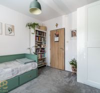 A room in a 3-room apartment with a bed, books, plants, and a lamp.