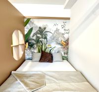 A bed with a patterned blanket in a studio apartment with tropical wallpaper and pillows.