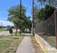The sidewalk in Bernolákovo runs alongside a garden fence, lined with trees and grass.