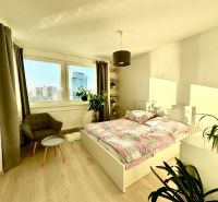 A bright bedroom with a bed, chair, and wood-decorated floor in a studio apartment.