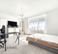 A bright room in a family house with a bed, a table, and a wooden-patterned floor.