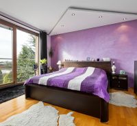 A bedroom in a family house with a purple wall, a bed, and a wooden decor floor.