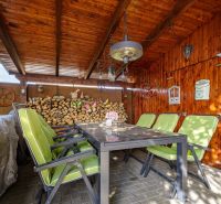 A wooden gazebo with a stone floor and green chairs by the family house.