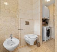 Bathroom with a toilet, bidet, and washing machine in a family house. Cream-colored tiles and flooring.