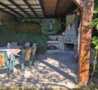 A covered terrace with a fireplace next to a cottage near Žilina, surrounded by greenery and seating.