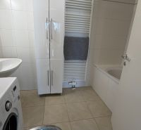 A bathroom in a 3-room apartment with a washing machine, sink, and bathtub on light tiles.