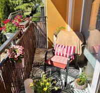 A balcony of a 3-room apartment with comfortable seating, flowers, and a view of greenery.