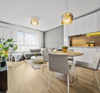 Living room in a two-room apartment with a wood-patterned floor, light furniture, and plants.