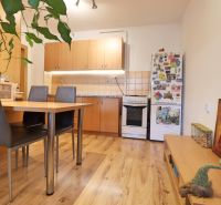 A kitchen in a 2-room apartment with a wooden decor floor, a dining table, and a refrigerator.