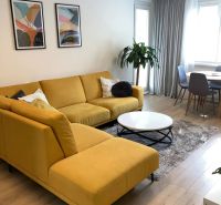 Living room of a 3-room apartment with a large yellow couch, wooden decor flooring, and a plant.