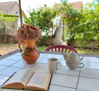 A covered table in the garden of a family house with a book, a teapot, and a flower vase.