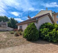 A family house in Matúškovo with a lawn, shrubs, and adjacent buildings under a blue sky.