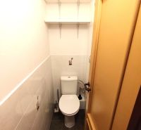 A compact toilet with hanging shelves and light tiles in a studio apartment.