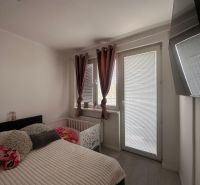 A bedroom in a 3-room apartment with a large window, curtains, and a TV on the wall.