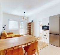 A dining room with a yellow sofa, a wooden table, and a kitchen with a stainless steel refrigerator.