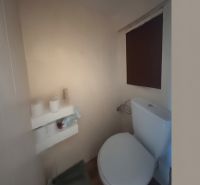 A bathroom in a 3-room apartment with a toilet, a shelf, and a blind on the window.