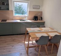 The cottage has a kitchen with wooden decor on the floor, a large window, and a dining table.
