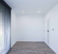 A room with white walls, dark blinds, and a wood-patterned floor in a family house.