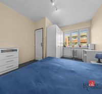 A room in a 3-room apartment with a bed, white cabinets, and windows with plants.