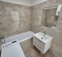 A bathroom in a studio apartment with a bathtub, sink, and washing machine; light stone tiles.