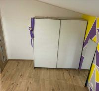 A room with a white wardrobe, a colorful wall, a wooden decor floor, and a toy.