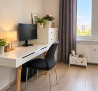 A workspace in a 3-room apartment with a desk, computer, and flowers on a floor with a wooden decor.