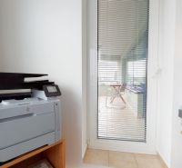 Printer on a wooden shelf, next to glass doors with vertical blinds.