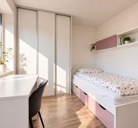 A bright room in a family house with a bed, a table, and shelves, with a wooden patterned floor.