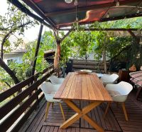 A terrace with a table and chairs under a pergola, surrounded by greenery and wooden boards.