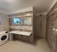 A bathroom in a family house with a washing machine, light tiles, and a shower corner.