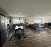 Open interior of a family house with a kitchen, dining table, sofa, and wooden decor flooring.