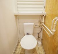 A small bathroom in a two-room apartment with a toilet, white walls, and pipes on the side wall.