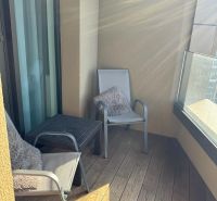 A balcony of a two-room apartment with a wooden decor floor, two chairs, and glass railings.