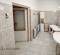 A bathroom in a two-room apartment with a washing machine, double sink, and cream-colored tiles.