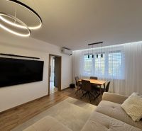 Living room in a 3-room apartment with a large television, wooden decor flooring, and a dining table.