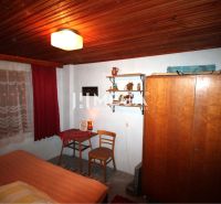 A cottage with a bed, chair, wardrobe, and shelf under a wooden ceiling.