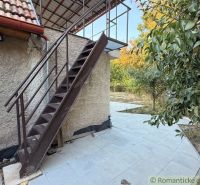 The exterior of a cottage in Nové Zámky with a metal staircase and a terrace surrounded by greenery.