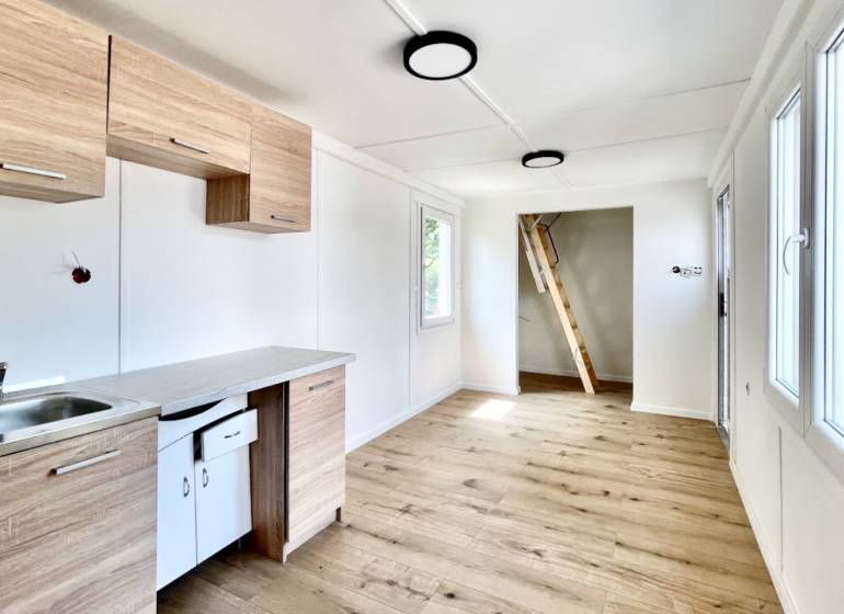 A kitchen in a cabin with a wooden floor, white walls, and a ladder to the upper floor.