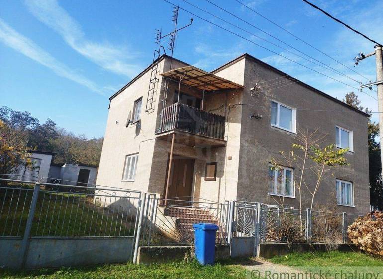 A two-story family house with a balcony, fencing, and a blue waste bin on a grassy plot.