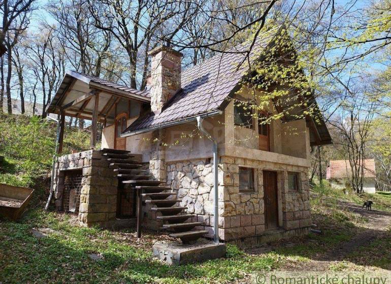 A stone cottage in the forest with an outdoor staircase and seating area, surrounded by trees in spring.