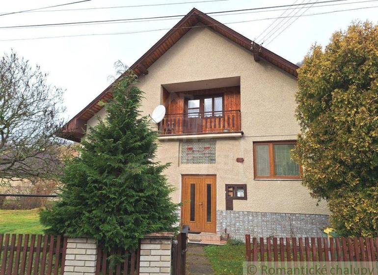 A family house with a wooden balcony, a satellite dish, and a garden behind a wooden fence.