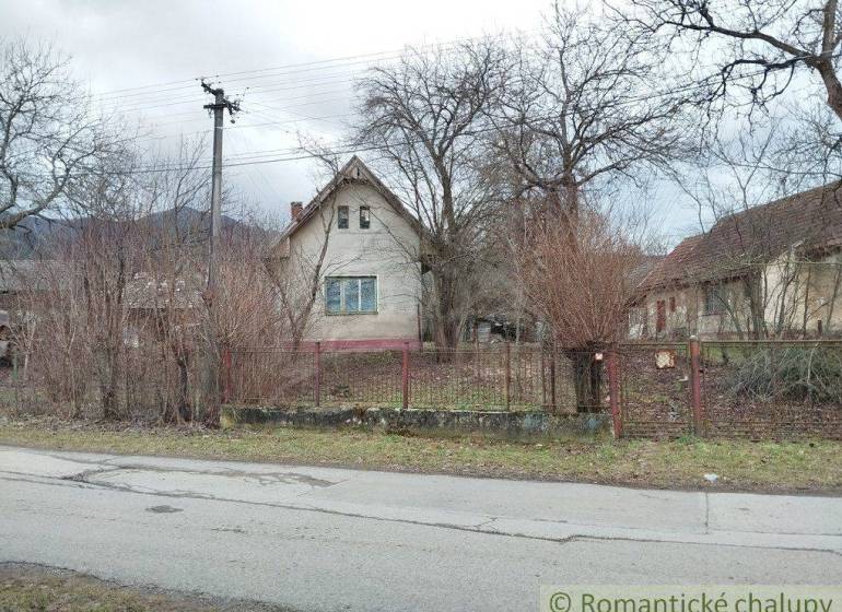 A family house surrounded by trees and bushes, a fence, a street, and a cloudy sky.