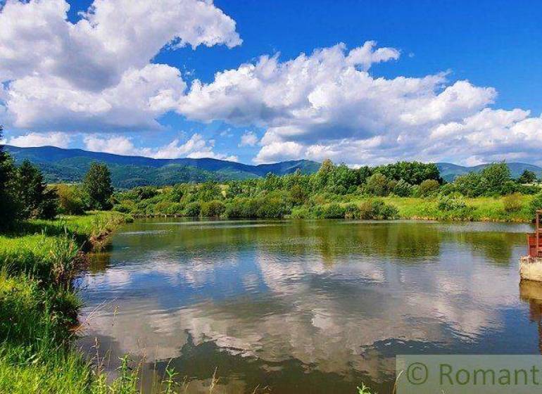 In the picture, there is a tranquil river with a bridge, surrounded by green hills and a blue sky. The area is suitable for plots and living.