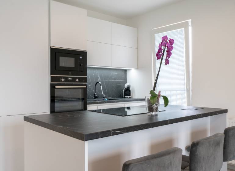 A kitchen in a 3-room apartment with built-in appliances, a black countertop, and a pink orchid.