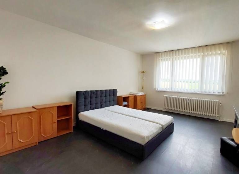 A simply furnished room with a bed, a dresser, and a large window in a studio apartment.