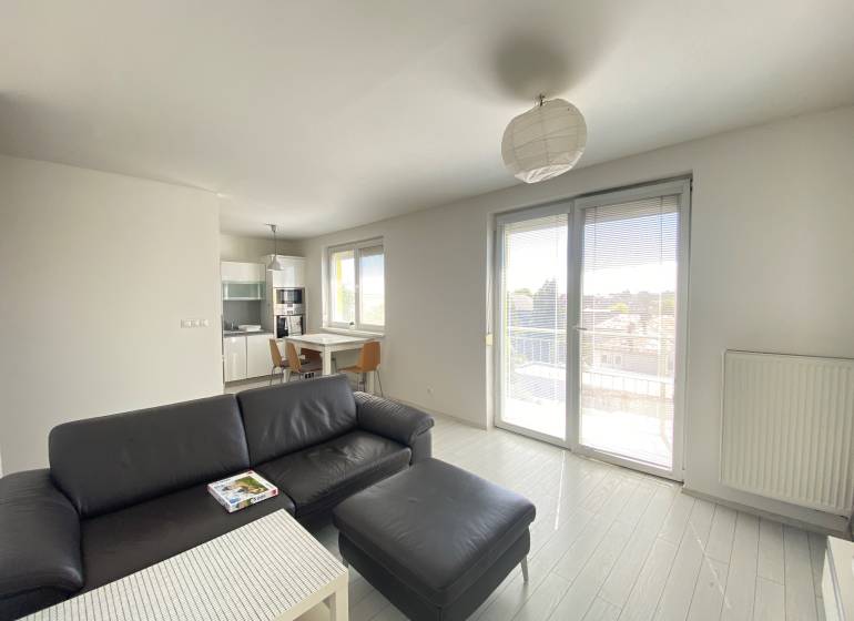 Living room and kitchen in a 3-room apartment with a black sofa, table, and balcony.